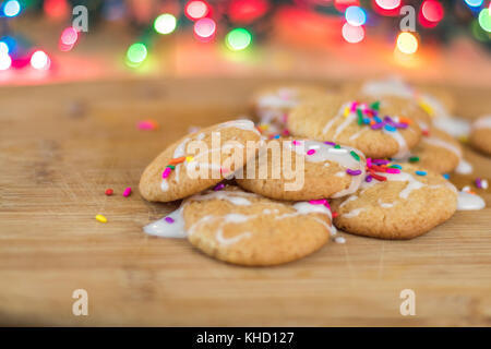 Frisch gebackene Sugar Cookies mit weißer Zuckerglasur und Rainbow farbige Streusel auf Holzbrett mit bunten Lichtern im Hintergrund, selektiver Fokus Stockfoto