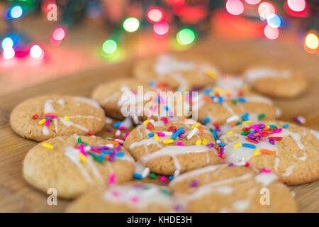 Frisch gebackene Sugar Cookies mit weißer Zuckerglasur und Rainbow farbige Streusel auf Holzbrett mit bunten Lichtern im Hintergrund, selektiver Fokus Stockfoto