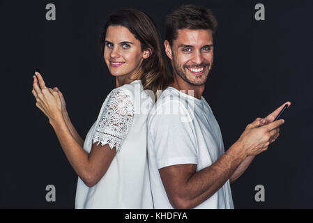 Paar Rücken an Rücken, beide halten Handys und fröhlich lächelnd Stockfoto