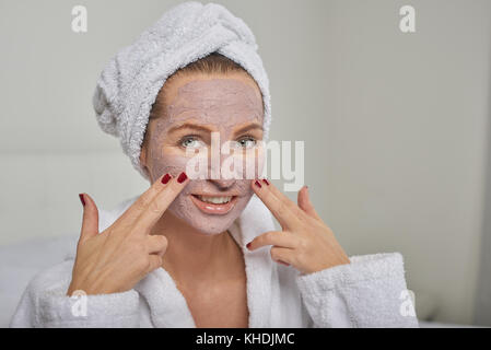 Attraktive Frau in einem weißen Bademantel Anwenden einer Maske, während in einem Handheld Spiegel zu betrachten, wie Sie sich mit einer Schönheitsbehandlung verwöhnen Stockfoto