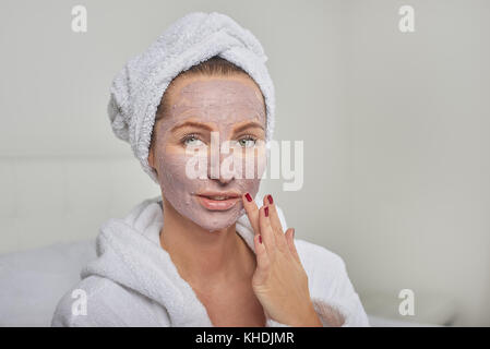 Attraktive Frau in einem weißen Bademantel Anwenden einer Maske, während in einem Handheld Spiegel zu betrachten, wie Sie sich mit einer Schönheitsbehandlung verwöhnen Stockfoto