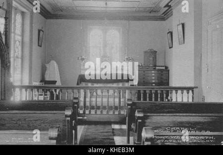 Ein altes Postkartenbild des Inneren der Mount Manresa Chapel, das einen direkten Vorderblick auf einen Altar mit einem Buntglasfenster auf der linken Seite, Holzgeländer auf der Vorderseite und einige Holzbänke im Vordergrund zeigt, Fort Wadsworth, Staten Island, New York, 1900. Aus der New York Public Library. Stockfoto