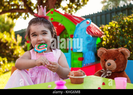 Baby Kleinkind Mädchen spielen Im freien Tea Party essen und Beißen eine große Lutscher mit bester Freund Teddy. rosa Kleid und Königin oder Prinzessin Krone. Stockfoto