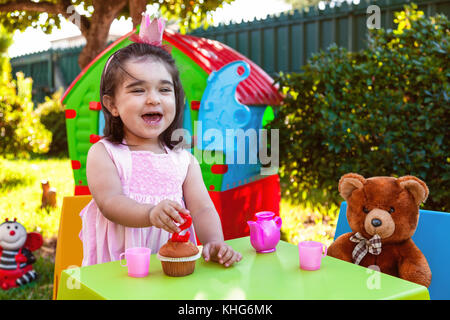 Glückliches lachendes Baby Kleinkind Mädchen im Freien zweiten Geburtstag holding Kerze. bester Freund Teddy, Playhouse und Tee. rosa Kleid und Krone Stockfoto