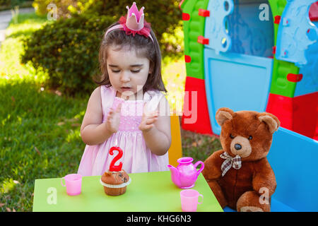 Baby Kleinkind Mädchen im Freien zweiten Geburtstag händeklatschen am Kuchen mit Teddy Bär als bester Freund, Playhouse und Kaffee. rosa Kleid und Krone. Stockfoto