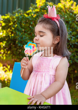 Happy Baby Kleinkind Mädchen riechen und genießen eine große bunte lollipop Geruch, Duft und Aroma in rosa Kleid als Prinzessin oder Königin mit Krone gekleidet Stockfoto