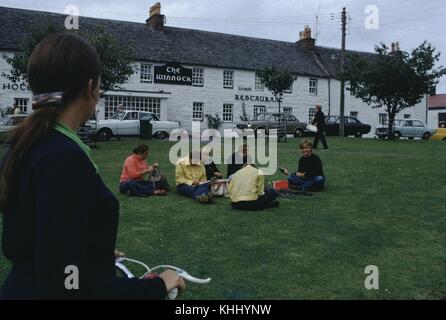 Gruppe junger Reisende, die auf dem Rasen vor dem Winnock Hotel and Restaurant sitzen, ein Mädchen mit einem Fahrrad, das mit dem Rücken zur Kamera im Vordergrund steht, Schottland, Großbritannien, 1966. Stockfoto