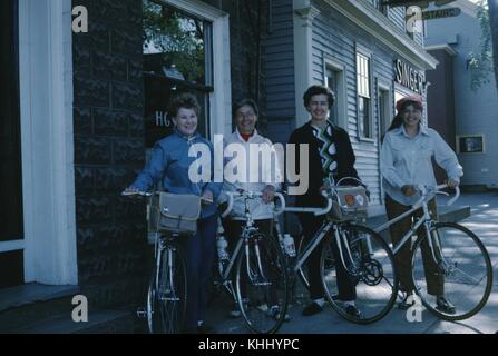 Vier Fahrradfahrerinnen posieren mit ihren Fahrrädern und lächeln, auf einem Bürgersteig vor einer Reihe von Geschäften, 1960. Stockfoto