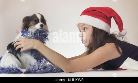 Schöne jugendlich Mädchen in Santa Claus hat und Hund continental Toy Spaniel Papillon in's neue Jahr Lametta freudig Spielen auf weißem Hintergrund Stockfoto