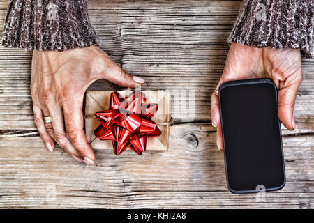 Nahaufnahme von einem Smartphone in den Händen einer Frau und einem Geschenkkarton mit einer roten Schleife auf einem Holztisch. freier Platz für Text. Kopieren Sie Raum. Blick von oben. Stockfoto