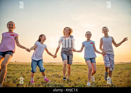 Mutter und 4 Kinder, die Spaß in das Feld, halten sich an den Händen und laufen in Richtung der Kamera im Freien, im Sommer Feld bei Sonnenuntergang. Sie legere sind c Stockfoto