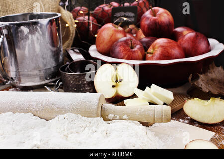 Alte hölzerne Rolling Pin bestäubt mit weißem Mehl über eine rustikale Tisch. Apfelkuchen Zutaten von Äpfeln und Butter mit Sichter und Messbecher im Hinterg Stockfoto