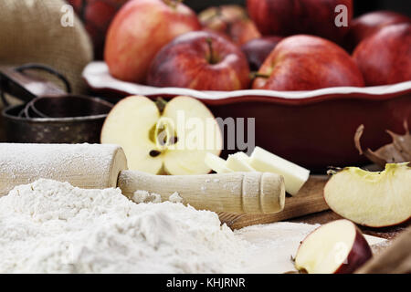 Alte hölzerne Rolling Pin bestäubt mit weißem Mehl über eine rustikale Tisch. Apfelkuchen Zutaten von Äpfeln und Butter im Hintergrund. Extrem flache Dept Stockfoto