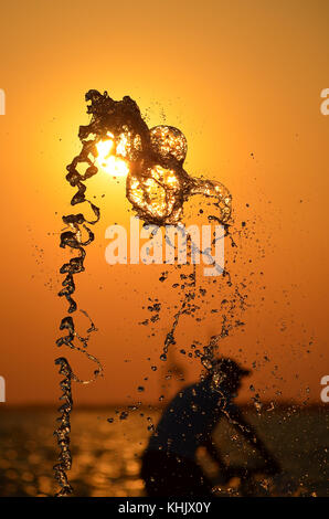 Wasser spritzen auf Sonnenuntergang farben Hintergrund mit Menschen und Krane Silhouetten isoliert. Stockfoto