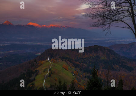 Jamnik, Slowenien - Blick Auf den wunderschönen goldenen Sonnenuntergang in der Jamnik St. Primoz Kirche. Julische Alpen im Sonnenlicht im Hintergrund Stockfoto