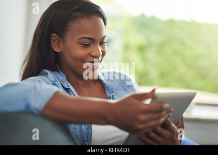 Lächelnden jungen afrikanischen Frau entspannend auf Ihr Wohnzimmer Sofa online surfen mit einem digitalen Tablet Stockfoto