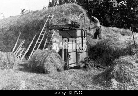 Ein Foto von zwei Bauern, die Heu mit einem handbetriebenen Bailer bailten, ein Mann steht auf einem Heuhaufen, mit einer Mistgabel, um es in den Bailer zu bewegen, der andere rollt einen fertigen Heuweg aus der Maschine, Das Foto wurde in einer Zeit des Übergangs von traditionellen Methoden zu moderneren, die Maschinen, Camargue, Frankreich, Juli 1922 aufgenommen. Stockfoto