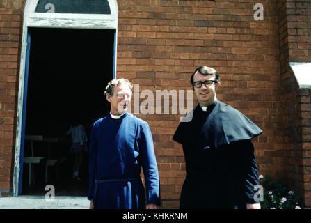 Ein Foto von zwei Priestern, die vor einer Kirche stehen, der Priester links trägt ein blaues Gewand, während der rechte ein schwarzes Gewand trägt, einfache Holzbänke und ein junges Mädchen in einem Rock sind in einer Tür zu der Kirche mit Backsteinmauern zu sehen, 1965. Stockfoto