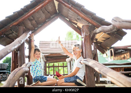 Bruder und Schwester sitzen einander gegenüber in einem Holzhaus am Spielplatz mit Sieg Geste Stockfoto