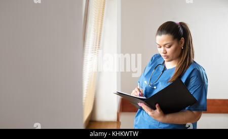 Junge weibliche spanischer Arzt lesen und Ausfüllen eines Formulars, stehen neben einem Fenster in einem Krankenhaus Flur Stockfoto