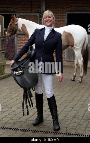 Attraktive blonde Frau mit ihrem Skewbald-Pferd in einem schwarzen Ledersattel. In einem Stableyard. November 2017 Stockfoto