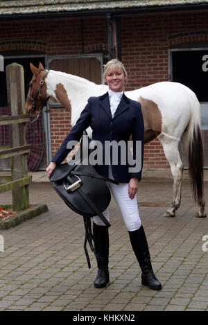 Attraktive blonde Frau mit ihrem Skewbald-Pferd in einem schwarzen Ledersattel. In einem Stableyard. November 2017 Stockfoto