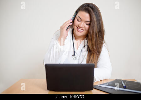 Glücklich lächelnde junge weibliche spanischer Arzt hinter dem Schreibtisch und sprechen auf dem Mobiltelefon Stockfoto