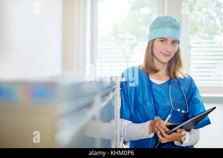 Junge weibliche Arzt in einer blauen Uniform steht drinnen in einer Klinik, mit Handy, Kugelschreiber und Ordner, in Richtung Aktenschrank lehnend, lo Stockfoto