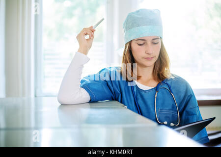 Junge weibliche nachdenklicher Arzt in einer blauen Uniform steht drinnen in einer Klinik, lehnte sich auf einer metallenen Aktenschrank, einem Stift und Suchen in Stockfoto