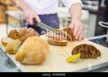 Die Hand eines männlichen Chef schneiden Vollkorn Brot mit Messer auf dem Tisch mit verschiedenen Arten von frisch gebackenem Brot. selektive Fokus, Platz für Text. Stockfoto