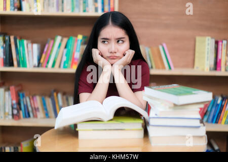 Asiatische Studentin langweilig lesen Buch an der Bibliothek mit einer Menge Bücher in der Universität. asiatische Student entmutigt lesen Buch für die Prüfung. Stockfoto