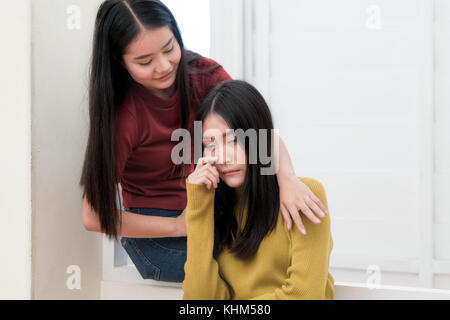 Schöne asiatische Frau umarmen depressiven Freund beim Sitzen auf Fenster zu Hause Stockfoto