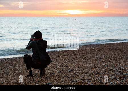 Brighton, UK. Eine junge Frau kauert auf dem Strand zu einem Foto auf den Sonnenuntergang. Stockfoto