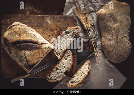 Ganze Laibe Brot und in Scheiben geschnitten auf rustikalen Holzbrett und Papier, mit Weizen Ohren und Messer, Ansicht von oben. Stockfoto