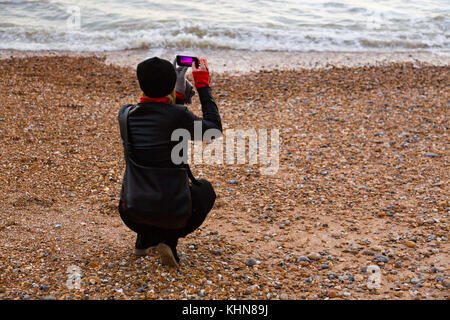 Brighton, UK. Eine junge Frau kauert auf dem Strand, ein Foto mit Ihrem Smartphone. Stockfoto