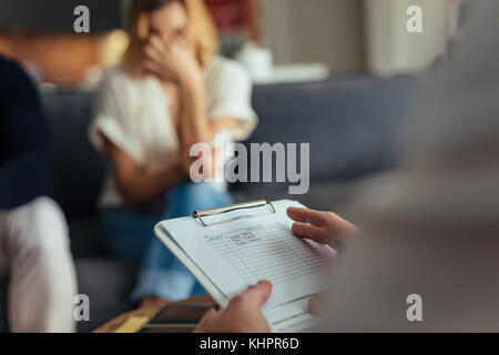 Konzentrieren Sie sich auf die Hände der männlichen Psychologen holding Zwischenablage. Traurig Paar sitzt auf einem Sofa im Hintergrund. Stockfoto