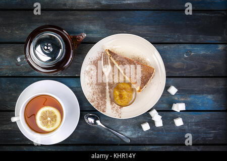 Stück Käsekuchen mit Marmelade und Tee Wasserkocher mit Zitrone auf einem farbigen Hintergrund Holz. nach oben anzeigen. Stockfoto