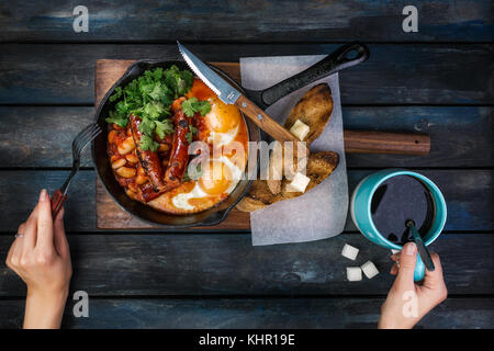 Frühstück in einer heißen Pfanne mit Spiegelei, Würstchen, Bohnen, Grün und Toasts. Frauen Hände mit Kaffee und Besteck. Auf einem farbigen Holz- Bac Stockfoto