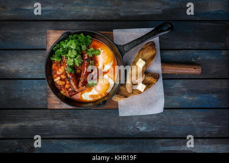 Frühstück in einer heißen Pfanne mit Spiegelei, Würstchen, Bohnen, Grün und Toasts. Auf einem farbigen Hintergrund Holz. nach oben anzeigen. Stockfoto