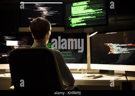 Arbeitsprozess im dim Büro: Programmiererin tragen weiße T-Shirt bis zur Programmierung gewickelt, beim Sitzen am Schreibtisch, Rückansicht Stockfoto