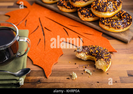 Mehrere Schokolade Donuts mit auf ein Fach eine Tasse Kaffee und eine Hälfte gegessen Donut gestapelt Streuseln. Herbst Tabelle einstellen, grün Serviette Stockfoto