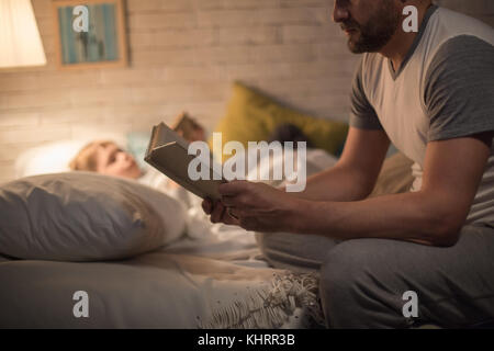 Seitenansicht Porträt des Menschen lesen Gute-Nacht-Geschichte zu kleinen Sohn sitzen am Rande von Bett im schwachen Licht mit unscharfen Form der Kind schlafen im Hintergrund. Stockfoto