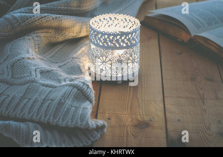 Kerzenlicht weißen Strickpullover offenes Buch auf der Plank Holz Tisch durch Fenster. Gemütliche winter Herbst Abend. Tageslicht authentische ruhige Atmosphäre. kin Stockfoto