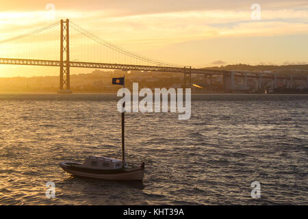 Golden leuchtet am späten Nachmittag des 25. April Brücke und einem kleinen Boot auf den Fluss Tagus. in Lissabon, Portugal. Stockfoto