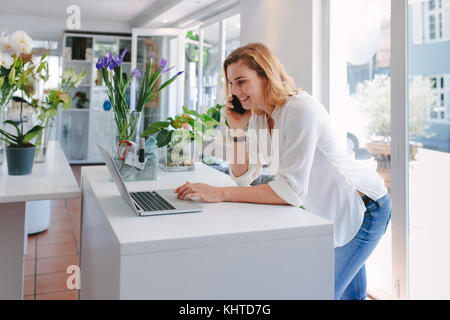 Weibliche florist Gespräch am Handy mit Laptop im Blumengeschäft. Glückliche junge Frau, die Reihenfolge, in der Gärtnerei. Stockfoto