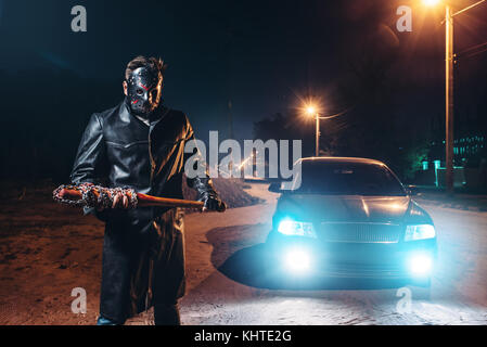 Serienmaniac in Ledermantel und Hockey-Maske, blutiger Baseballschläger in Metallkette in Händen gewickelt, gegen schwarzes Auto mit Licht in der Nacht. Horror, Stockfoto