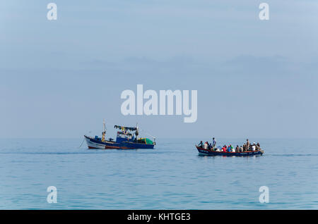 LUCIRA, ANGOLA - 17. MAI 2014: Kleine gemalte Fischerboot und kleineren Boot mit vielen Menschen am Atlantik Stockfoto