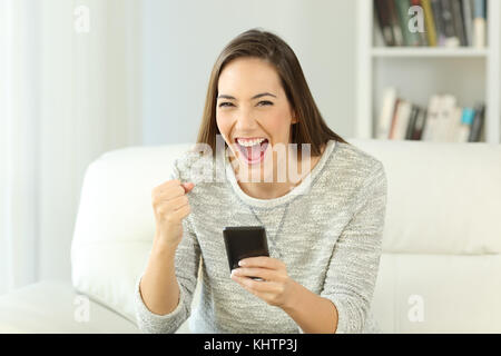 Vorderansicht Portrait von eine aufgeregte Frau mit Telefon, Sie sitzen auf einem Sofa im Wohnzimmer eines Hauses Innenraum Stockfoto