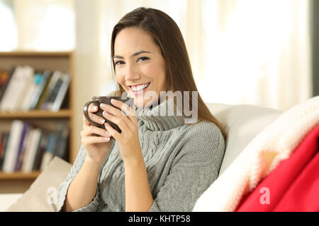 Portrait eines glücklichen Frau posiert Holding eine Kaffeetasse sitzt auf einem Sofa im Wohnzimmer in einem Haus im Innenbereich im Winter Stockfoto