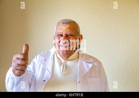 Alte Doktor mit Flecken auf der Stirn lächelnd und mit Daumen hoch Stockfoto